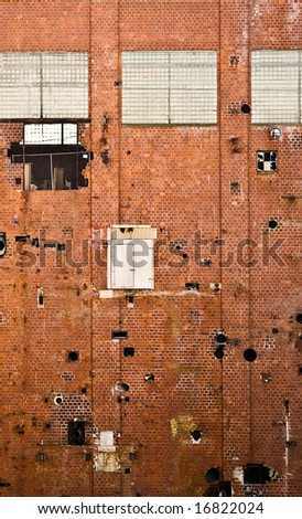 Side of old red brick building in factory demolition site. The building is full of holes, broken windows and an odd door several stories up with no stairs.