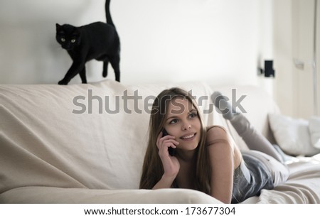 Blonde girl and black cat stretched on the couch talking on mobile phone