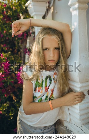 The portrait young girl about 9-12 years old with the blonde and loose hair that stands near the columns with the purple flowers in the rays of the setting sun. She wears top-shirt and shorts.