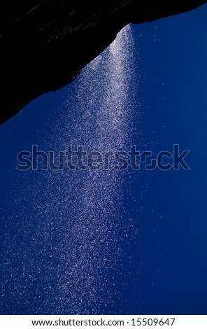 Backlit waterfall from rock cliff, sparkling water drops over blue sky