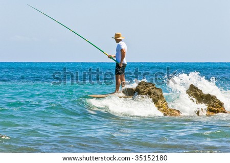 The fisherman in a vest and a hat fishing on stones in sea waves