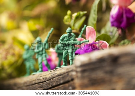 Green plastic toy soldier army unit running on top of an old weathered railway sleeper. Selective focus and wooden textured background and purple flowers