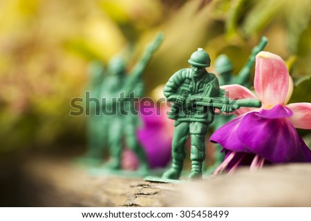 Green plastic toy soldier army unit running on top of an old weathered railway sleeper. Selective focus and wooden textured background and purple flowers