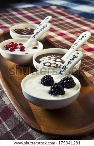 Closeup cream dessert with chocolate and berries in white porcelain bowls on wooden plate with ceramics measuring spoons. Checkered tablecloth.