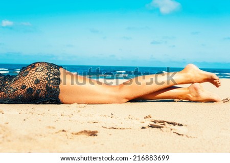 Luxuriant tanned young woman lying body and long legs in transparent reticulated dress on the beach sand. Outdoors.