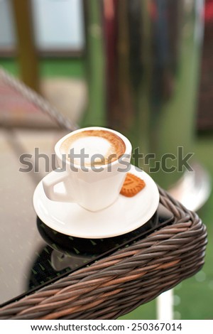 White porcelain cup of black coffee with whipped milk on rattan table in summer cafe. Outdoors closeup.