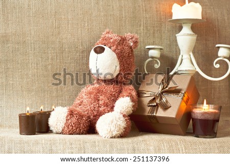 Romantic present. Teddy bear toy with gift box and candles on sack/burlap background. Filtered image. Indoors still-life.