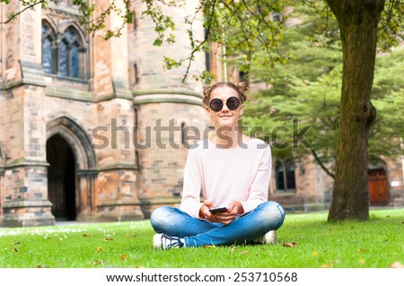 Young lady with mobile phone sitting on the grass in Glasgow University garden looking at camera. Summertime outdoors.