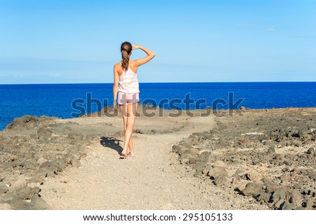 Young beautiful tanned teenage girl in pink shorts walking on Atlantic ocean rock coast. Tenerife, Canary islands, Spain. Multicolored summertime outdoors image on a blue sky background.