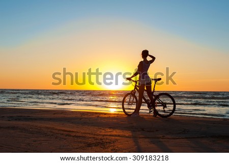 Moment in time. Young sporty woman cyclist silhouette contemplating the sunset on blue sky background on the beach. Summertime multicolored outdoors horizontal image.