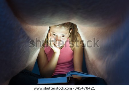 Young teenage girl hiding under blanket and enrapt reading interesting book at nighttime. Key light coming from book. Indoors horizontal image.