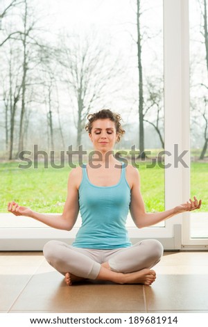 Beautiful young woman doing yoga exercise in her house in front of the big window with a nature view