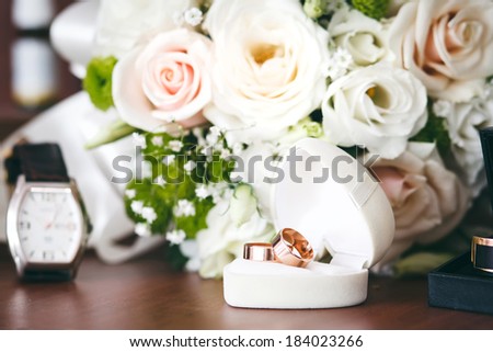 Gold wedding rings in white gift box in shape of heart with wedding bouquet and watch in background