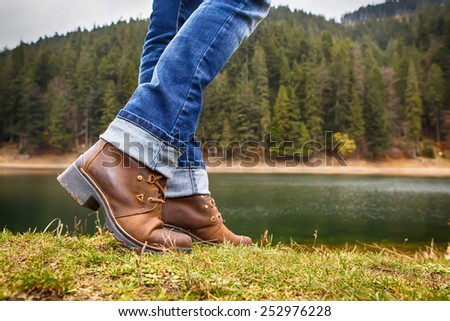 Woman in boots and jeans walks along the lake shore in autumn forest