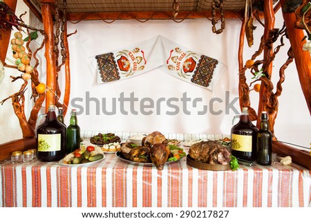 Grilled pig, smoked ham, bottles of wine and other foods on welcoming table, decorated in Ukrainian traditional style, at party in restaurant. Towel hangs on wall with inscription \
