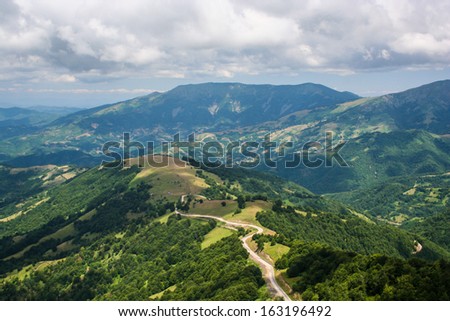 Road through high mountain range. Areal view on road going on top of the hill through high mountains with green forests on sides and dramatic rainy gray clouds in the sky.