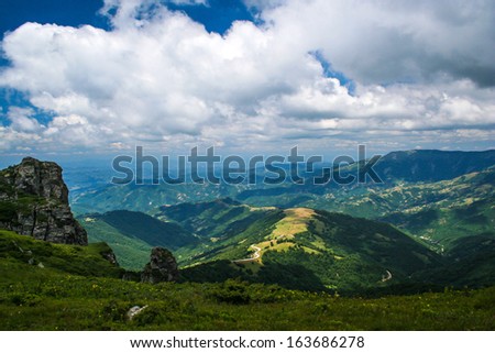 Road through high mountain range. Areal view on road going on top of the hill through high mountains with green forests on sides and dramatic rainy gray clouds in the sky.