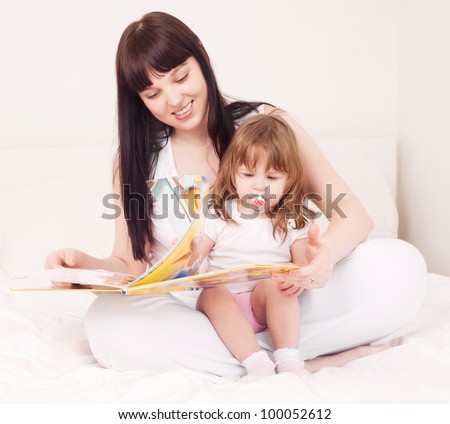 young mother and her one year old daughter reading a book in bed at home