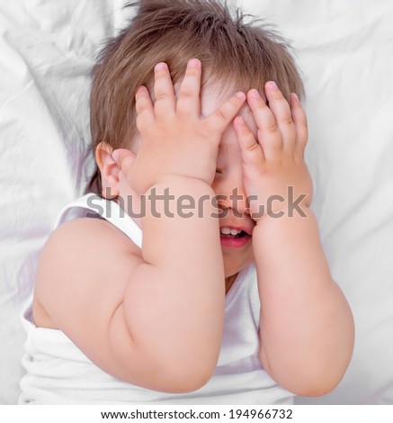 scared baby closing his face with hands, in bed at home, top view