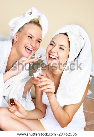 tow happy laughing girls sitting on the bed and applying nail polish