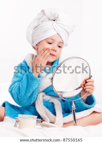 beautiful five year old girl with a mirror applying cream on her face on the bed at home