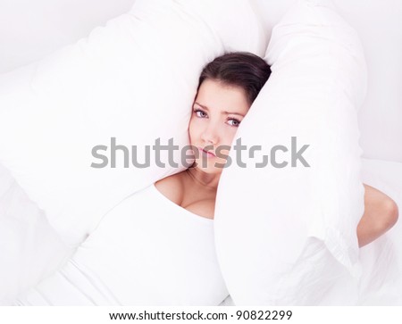 displeased  young brunette woman frowning and closing her ears with pillows, in bed at home