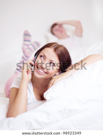 happy young woman talking on the phone and  sleeping man in bed at home (focus on the woman)