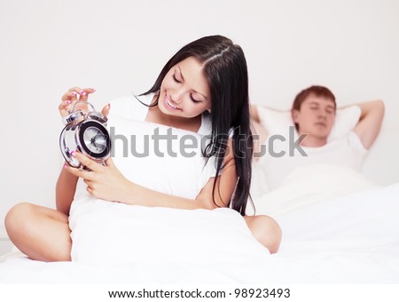 happy young woman with alarm clock and  sleeping man in bed at home (focus on the woman)