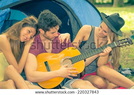friends on camping. A boy with guitar plays romantic song and the two girls on his side are very impressed