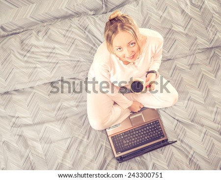 Young woman sitting on bed with a cup of coffee and computer - Woman having breakfast in bed and looking news at computer laptop