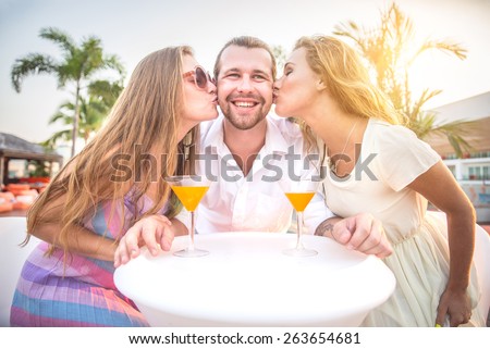 Two beautiful women kissing on cheeks a man - Friends at party drinking cocktails and having fun - Three tourists drinking aperitif in a tropical luxurious restaurant
