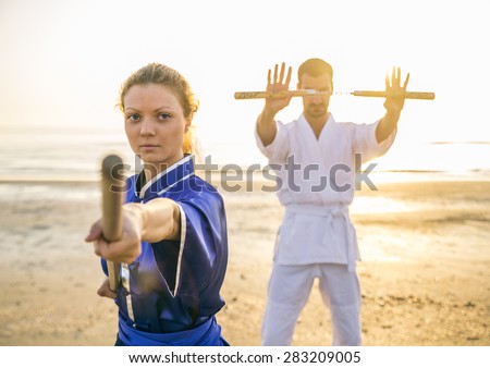 Two martial arts athletes training with weapons on the beach at sunrise - Sportive man and woman with stick and nunchaku - Kung fu and karate masters