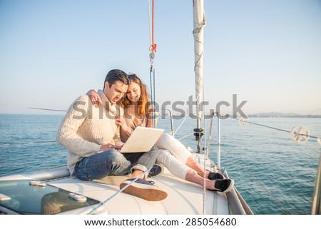 Couple sitting on a sailing boat looking at laptop computer - Beautiful woman and attractive man having fun on a boat while on vacation