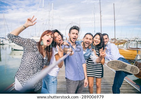 Best friends taking a self portrait with selfie stick - Group of young and happy people on vacation at harbor with sailing boats in the background