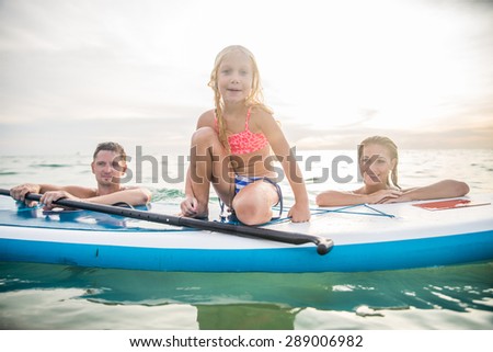 Happy family with paddle board swimming in the ocean - Pretty young girl smiling while learning to paddle - Portrait of active and sportive family on vacation on summertime