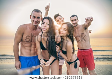 Multiethnic group of friends enjoying vacation  and taking a picture with selfie stick - Mixed group of several people having fun on the beach at sunset