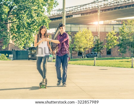 Young playful couple having a romantic date outdoors - Sportive man teaching her girlfriend to skateboarding - Teen lovers spending time together and having fun