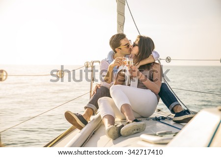 Beautiful couple of lovers sailing on a boat - Couple kissing and holding champagne to celebrate - Two fashion models posing on a sailing boat at sunset