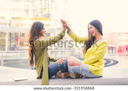 Two beautiful young women giving high five - Pretty girls sitting on a bench outdoors and having fun - Best girlfriends making a promise