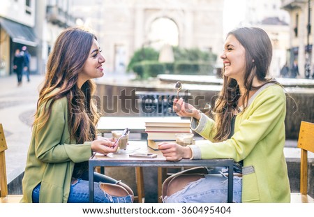 Two pretty girlfriends laughing while sitting in a bar outdoors - Students having pause and drinking cappuccino - Best friends talking and having fun