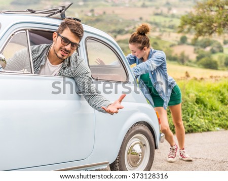 Engine break down.Strong young woman pushing a vintage car while man is emboldening her.Transportation, teamwork, funny concept