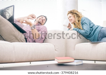 Cheerful couple lying on the couch and looking at laptop computer - Partners talking and smiling while relaxing in the living room, domestic life scene