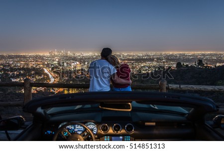 Couple enjoying skyline view from their car in the night. Focus on the Los angeles city in the background