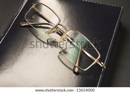 Glasses with gold-rimmed is lying on black leather an organizer