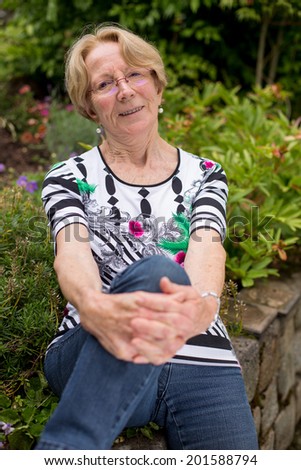 A nice elderly woman sits in the garden and smiles into the camera bending one knee and leaning on it