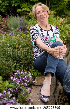 A nice elderly woman smiles sits in the garden and smiles into the camera bending one knee and leaning on it