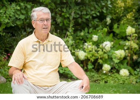 An elderly man is sitting relaxed in a garden while resting his arms on his knees