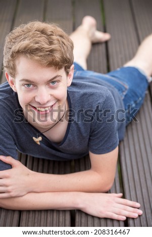 A young blonde male model is lying on the ground, resting on his arms and smiling into the camera