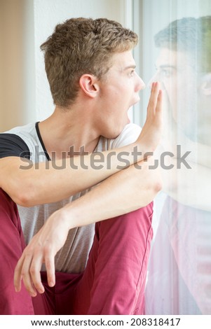 A young male model is pressing his nose against his own reflection in a window and touching it with his hands. He is angry!