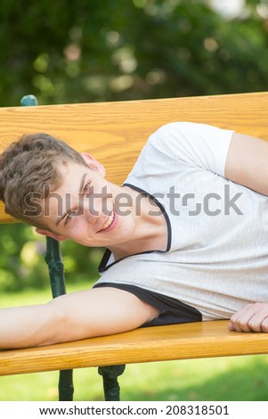 A young male model is smiling at the camera and laying relaxedly on a beautiful wooden bench in a green garden with his arm laying on the bench